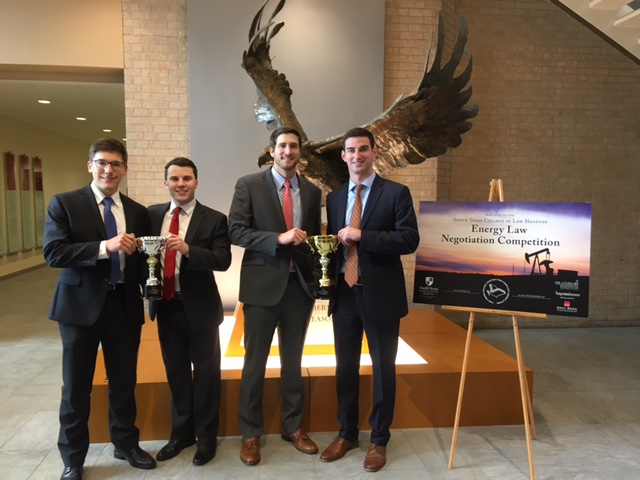 Four young men in business suits hold up trophies. In the background, a statue of an eagle spreading its wings.